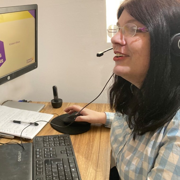 A woman with dark hair and glasses sits at a desk with a monitor and headset.